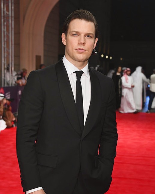 Jake Lacy posing in a white shirt and black suit.